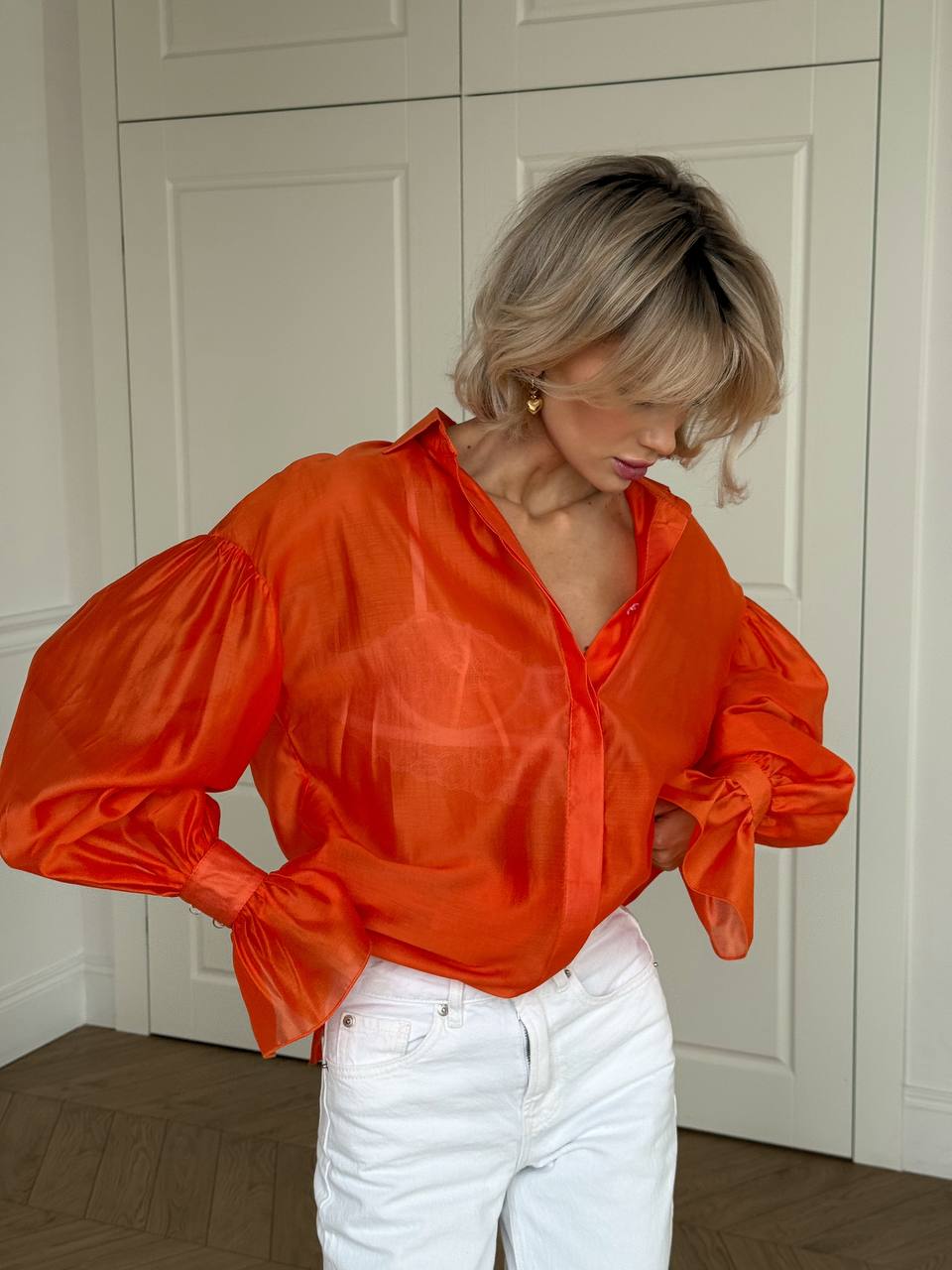 Woman wearing an orange blouse with puffed sleeves and white pants in a room.