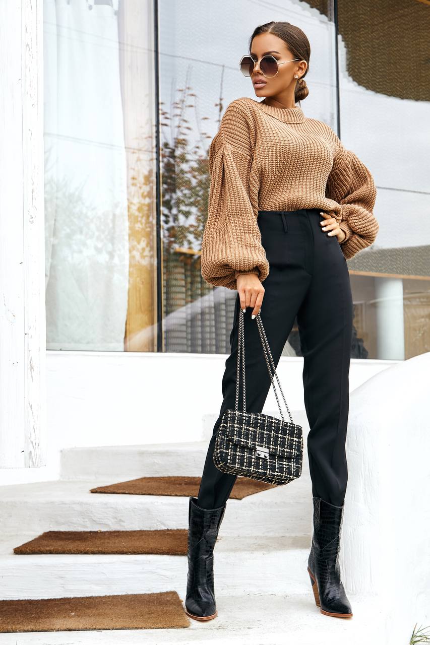 Woman in a tan sweater and black pants holding a handbag, standing on steps.