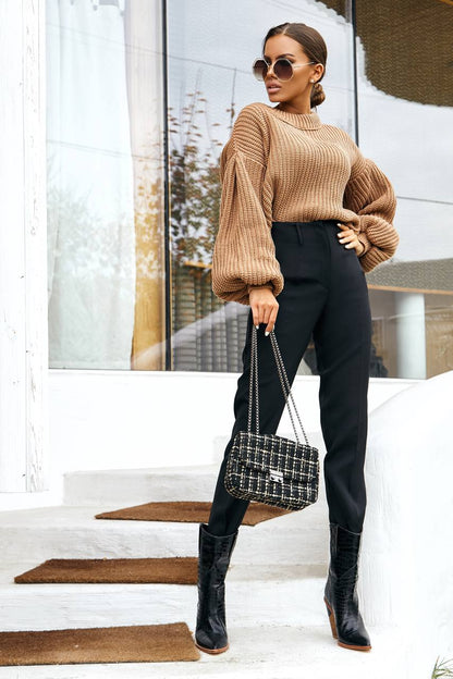 Woman in a tan sweater and black pants holding a handbag, standing on steps.