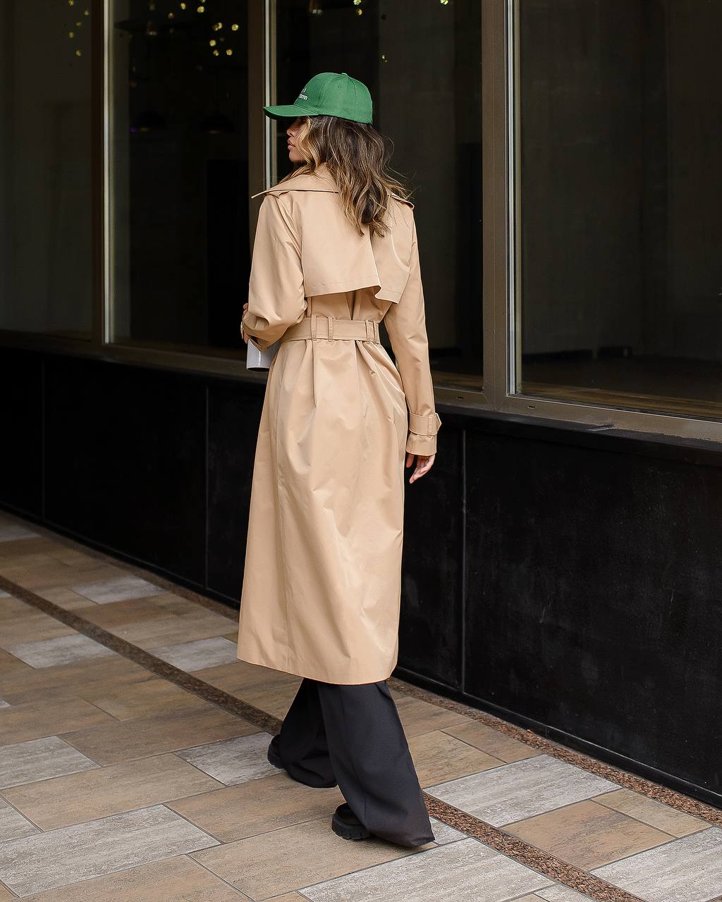 Person wearing a beige trench coat and green cap standing on a sidewalk.