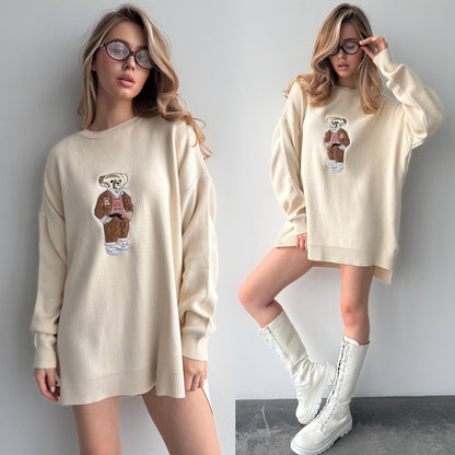 Woman wearing a cream-colored sweater with a bear graphic, paired with white boots against a plain background.