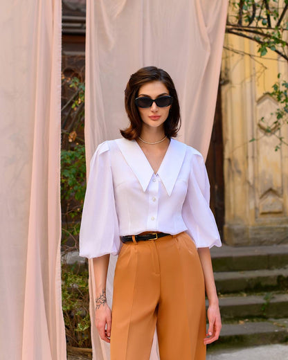 Woman wearing a white blouse and brown pants standing in front of beige curtains.
