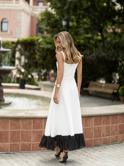 A woman wearing a white sleeveless midi dress with a black ruffled hem, standing outdoors near a fountain and greenery.
