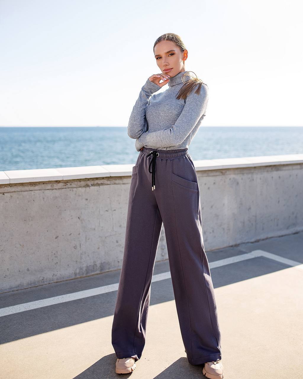 Woman in a gray sweater and dark pants standing by a waterfront.