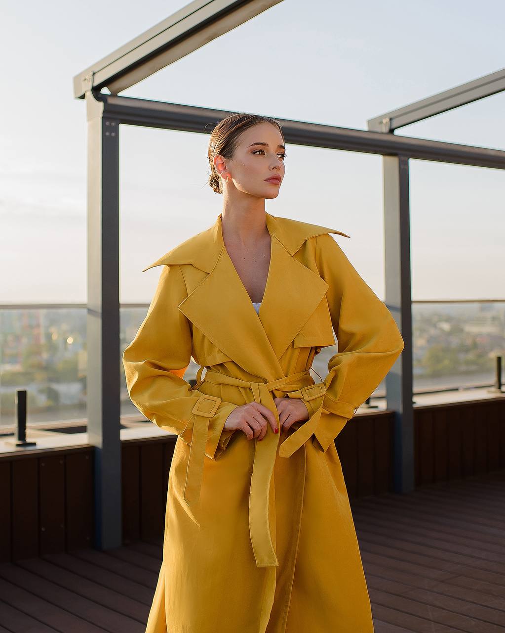 Woman wearing a yellow trench coat on a rooftop with a cityscape view.