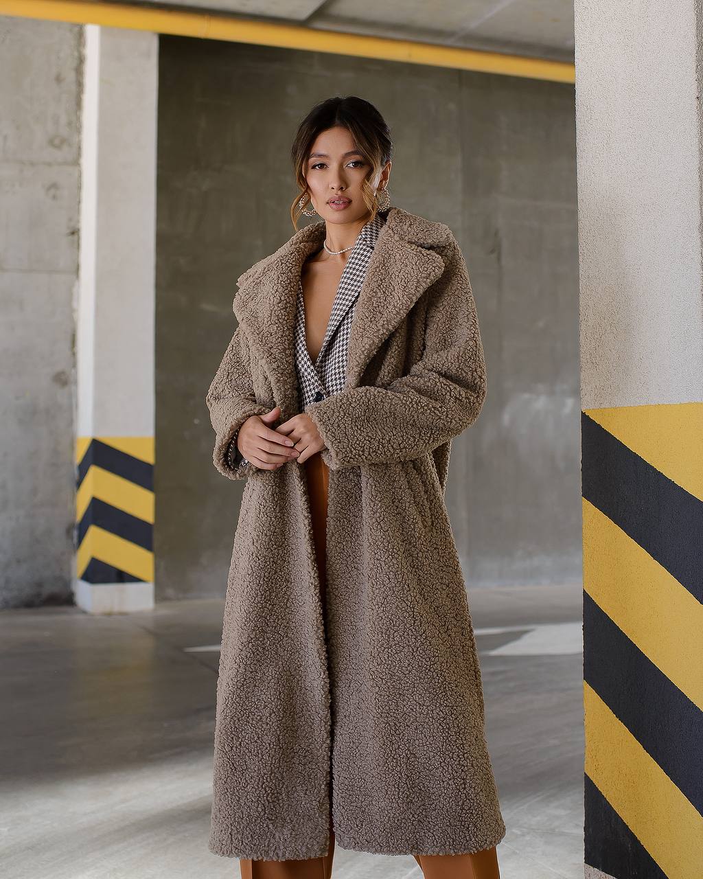Woman wearing a long brown coat in an industrial setting with concrete walls and yellow and black stripes.