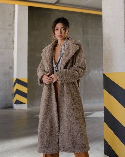 Woman wearing a long brown coat in an industrial setting with concrete walls and yellow and black stripes.