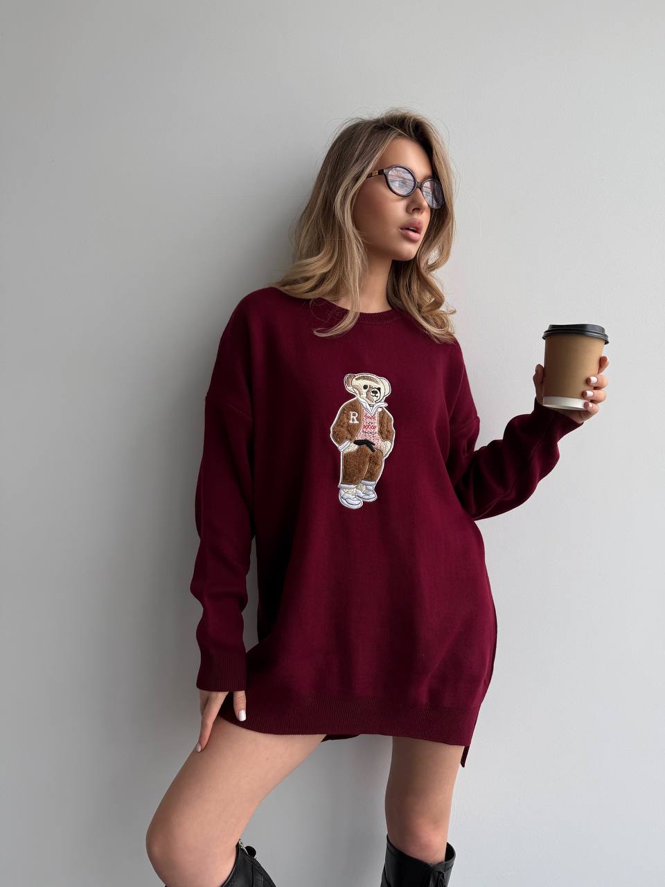 Woman wearing a burgundy sweatshirt with a bear graphic, holding a coffee cup against a plain background