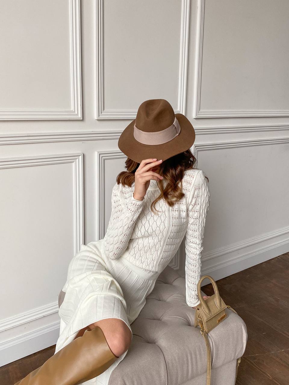 Woman in a white dress and brown hat sitting on a chair against a gray paneled wall.