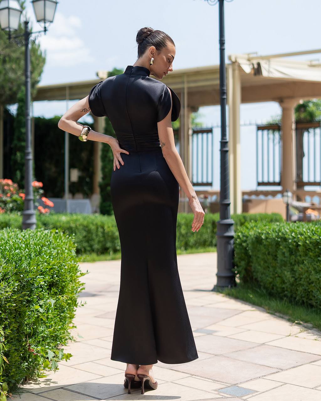 Woman in a black dress standing on a paved path with greenery and a building in the background