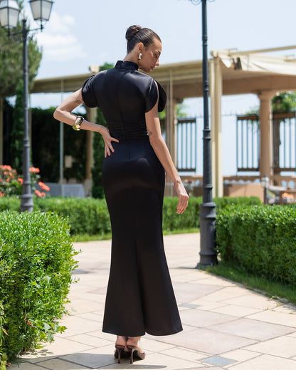 Woman in a black dress standing on a paved path with greenery and a building in the background
