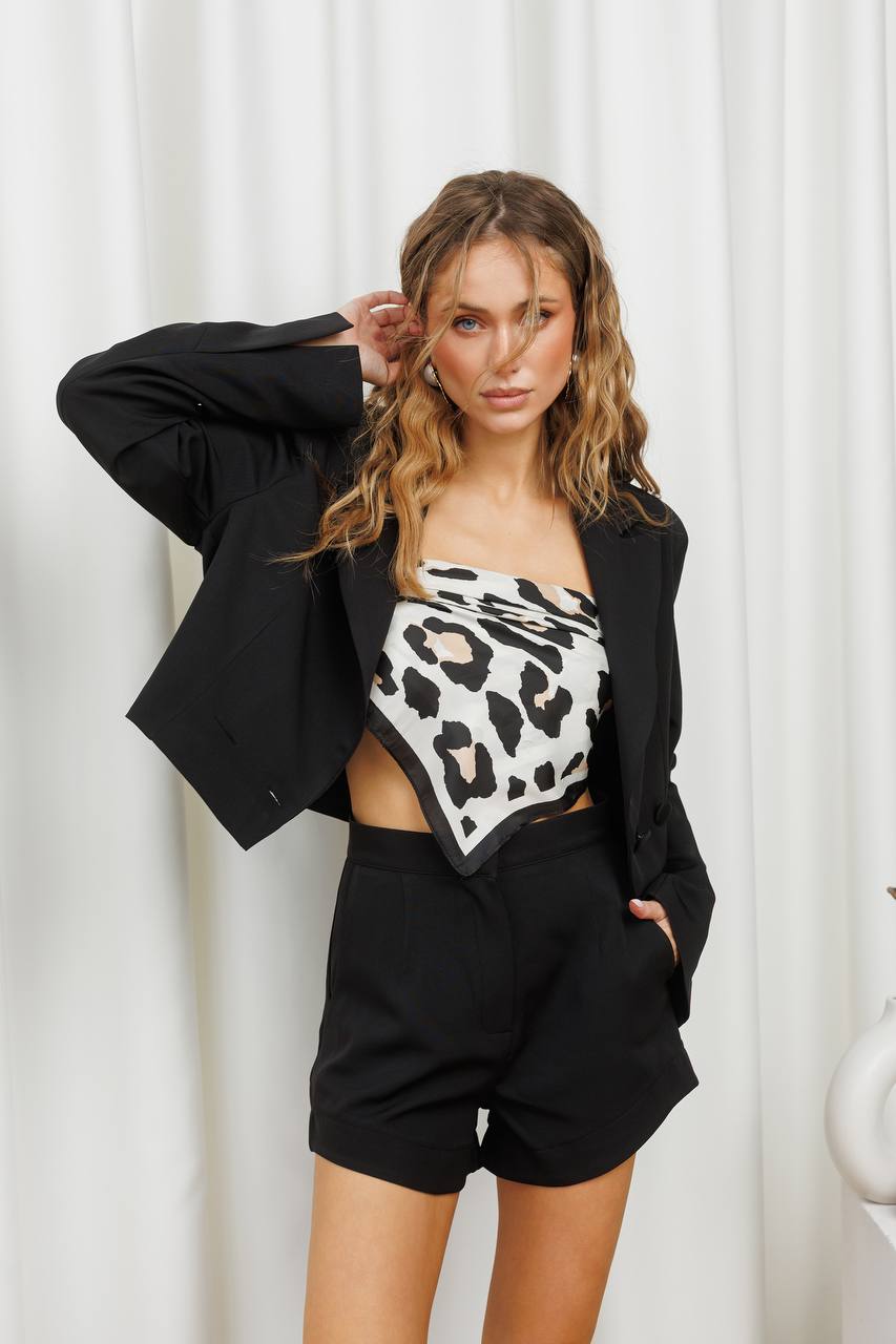Woman wearing a black blazer over a leopard print top with high-waisted black shorts against a white curtain background.