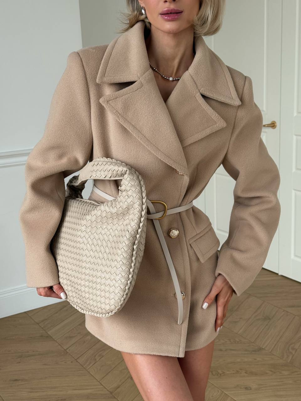 Woman wearing a beige coat holding a woven handbag indoors.