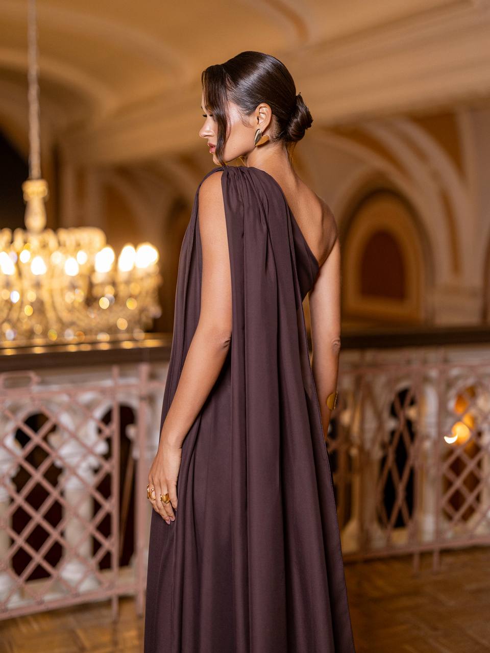 Woman in a brown dress standing in an elegant indoor setting with chandeliers.