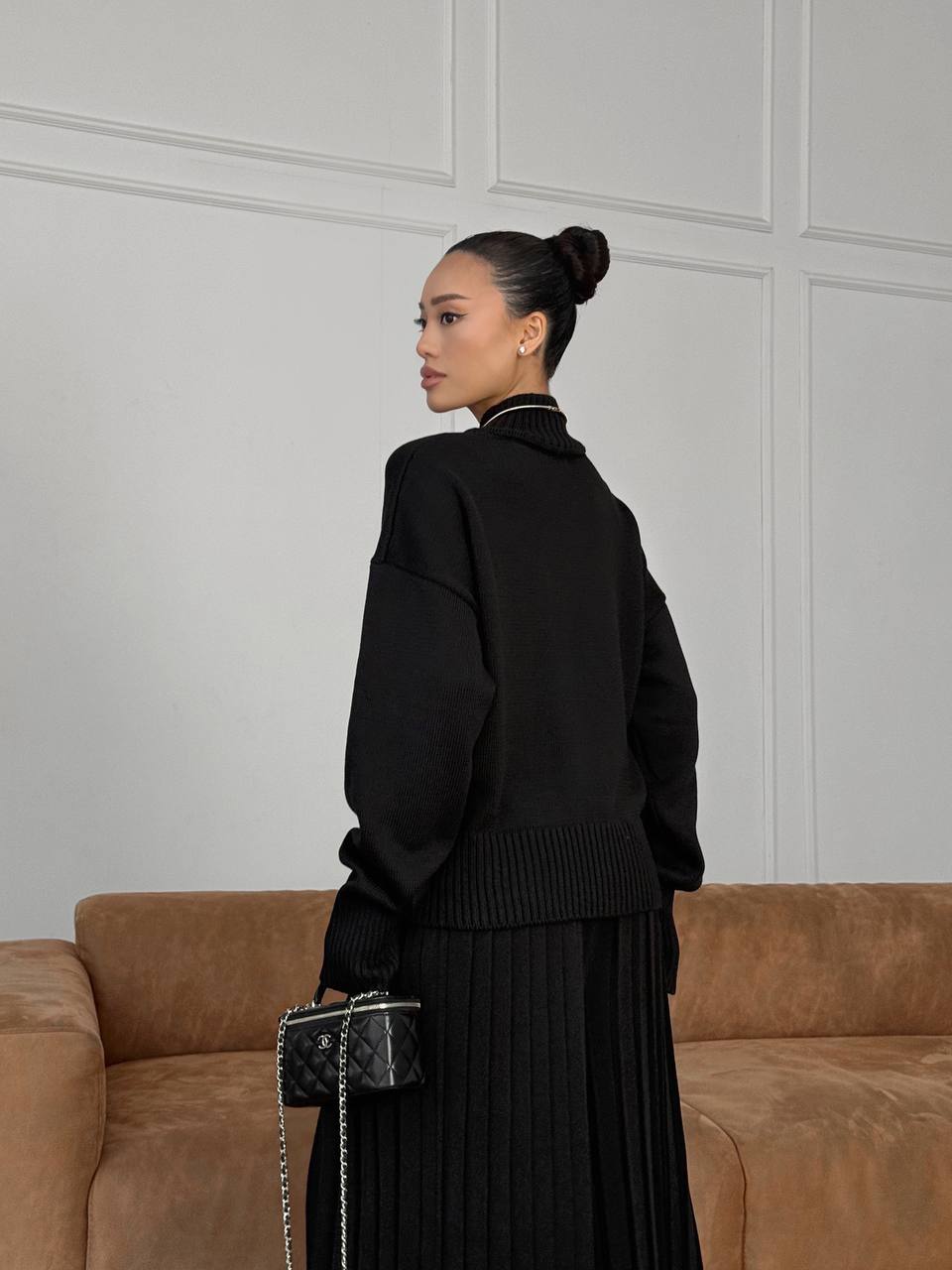 Woman in a black outfit standing in front of a brown sofa with a neutral wall background