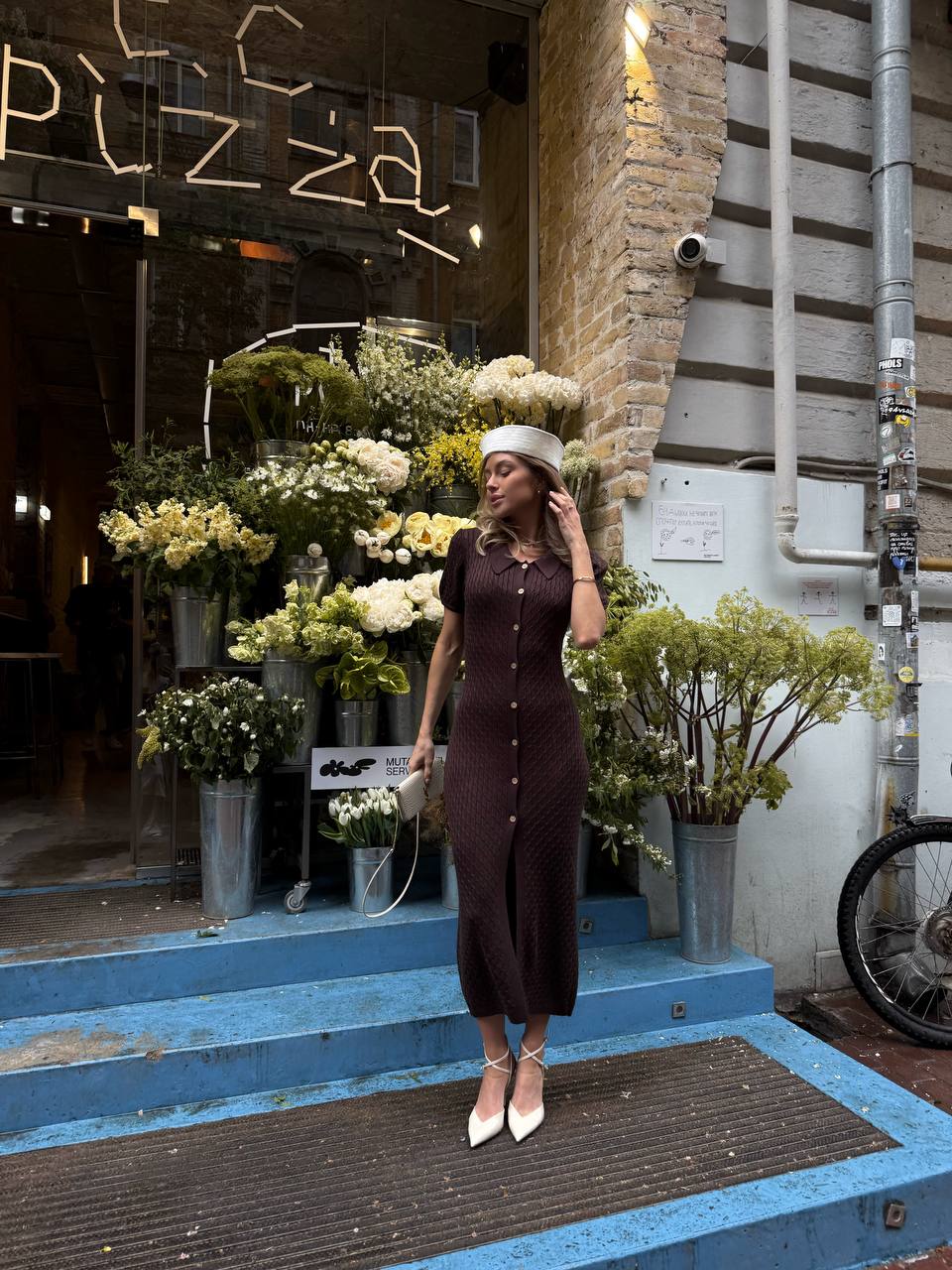 Woman in a brown dress standing in front of a pizza restaurant with flower arrangements.