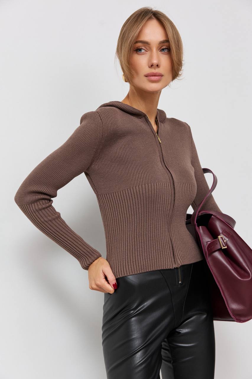 Woman wearing a brown sweater with a hood and black leather pants, holding a purple handbag on a white background.