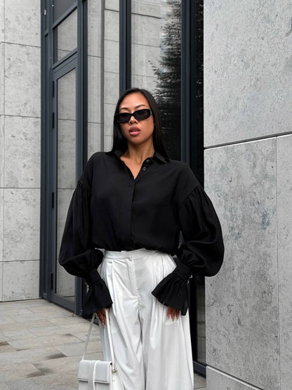 Woman in black blouse and white pants standing against a gray building.
