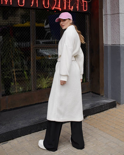 Person wearing a long white coat and black pants standing on a street with a neon sign in the background.