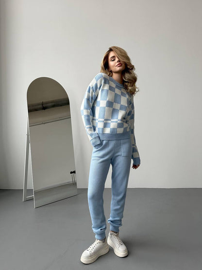 Woman wearing a blue checkered sweater and pants standing in front of a mirror.