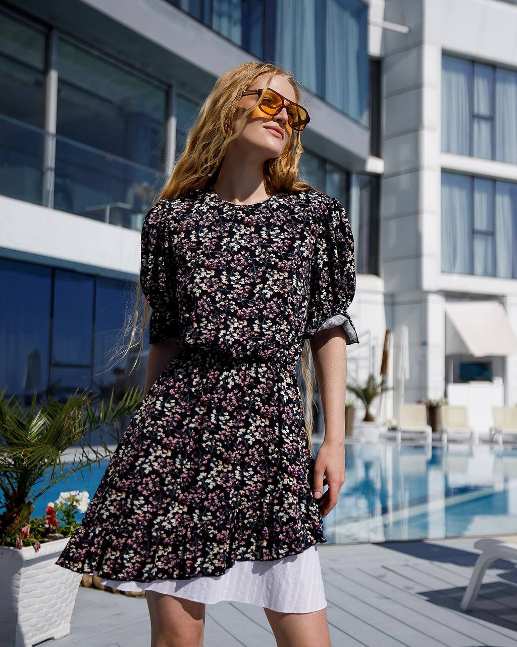 Woman wearing a floral dress and sunglasses by a poolside building.