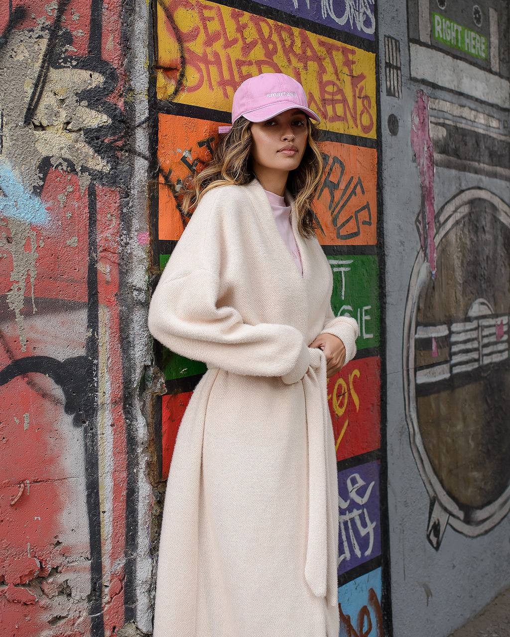 Woman in a long coat and pink cap standing against a graffiti-covered wall.