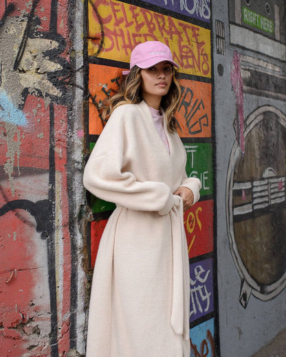 Woman in a long coat and pink cap standing against a graffiti-covered wall.