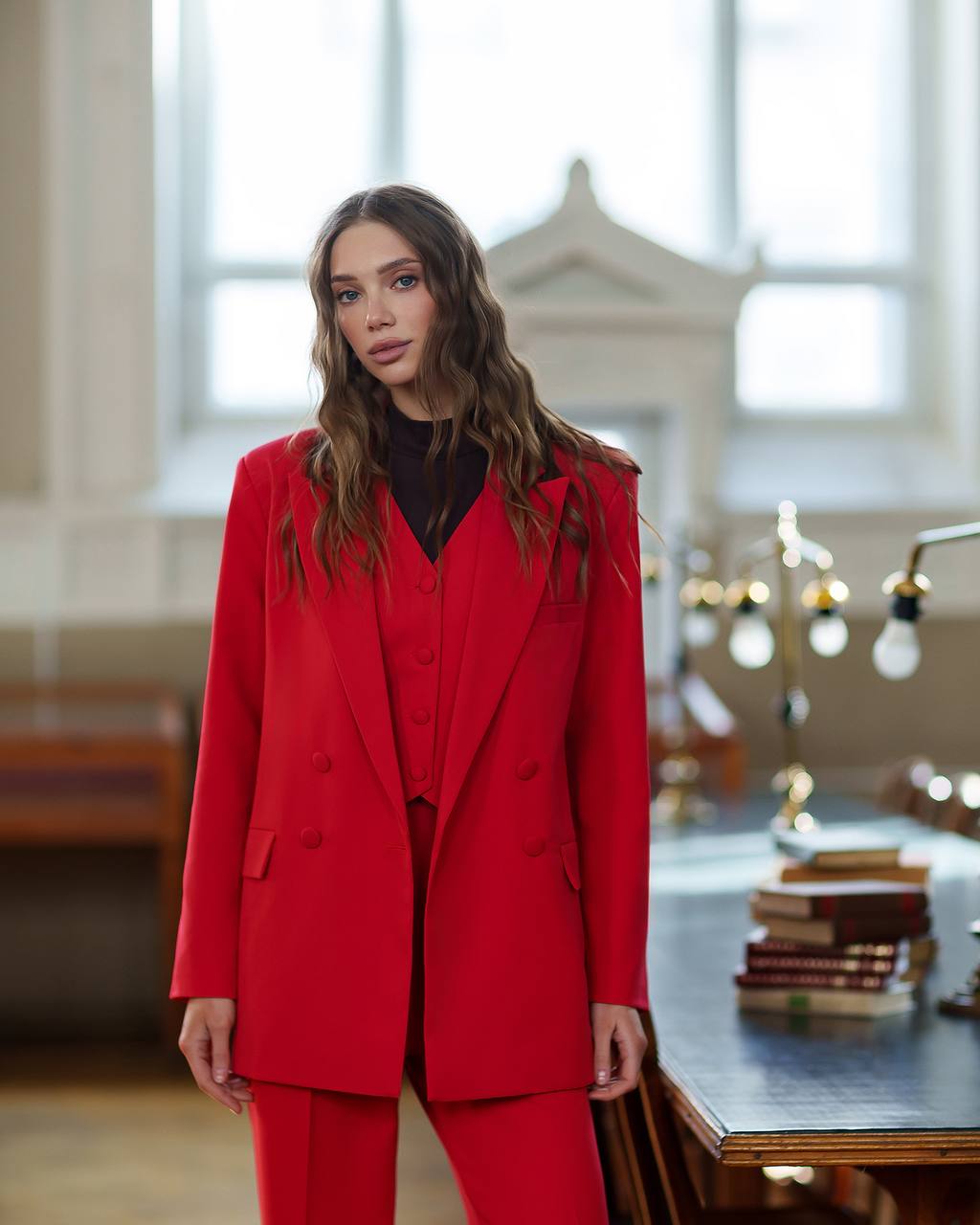 Woman wearing a red suit standing in an indoor setting with large windows.