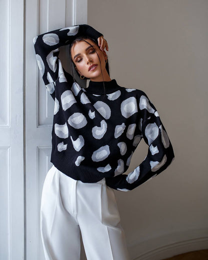 Person wearing a black sweater with white abstract patterns and white pants against a neutral wall.