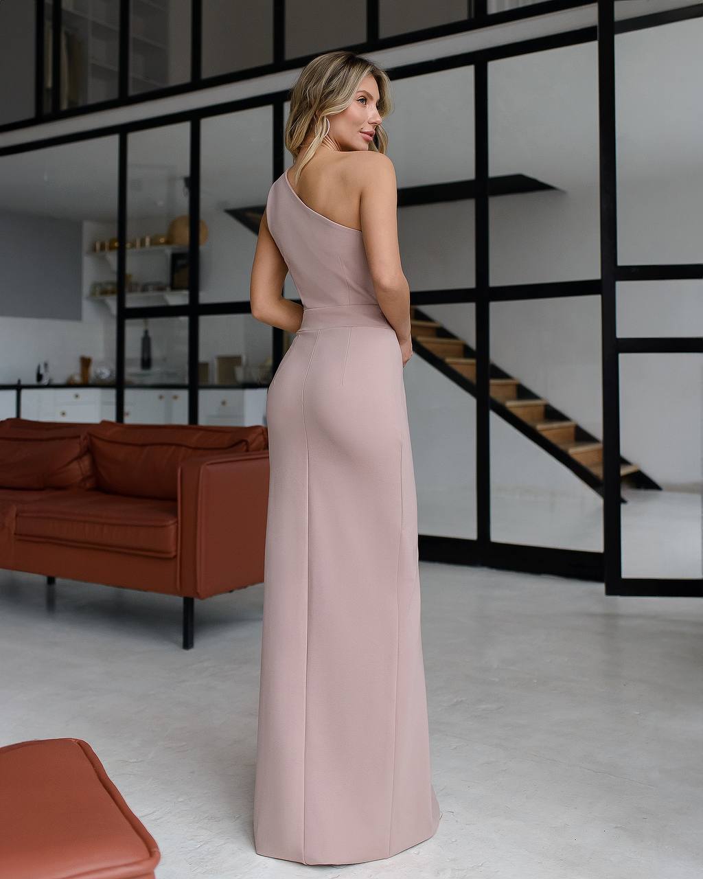 Woman in a pink dress standing in a modern interior setting with a brown sofa and glass walls.
