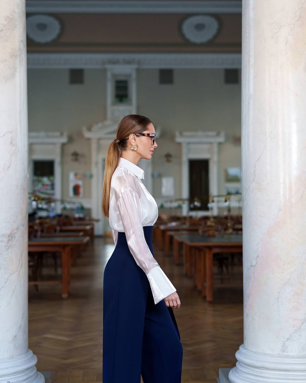 Woman in a white blouse and navy pants standing between two columns in an indoor setting.
