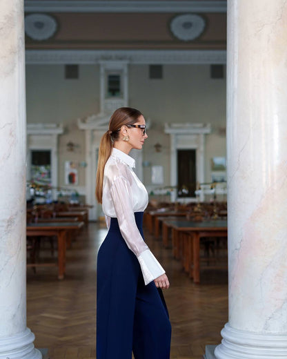 Woman in a white blouse and navy pants standing between two columns in an indoor setting.