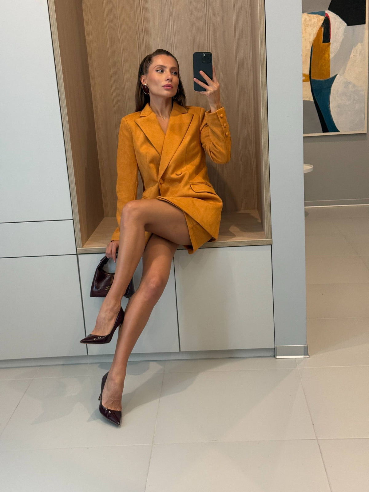 A woman takes a mirror selfie wearing a mustard yellow tailored blazer dress with structured shoulders and a wrap-style front, paired with dark pointed heels, seated on a built-in bench in a modern interior.