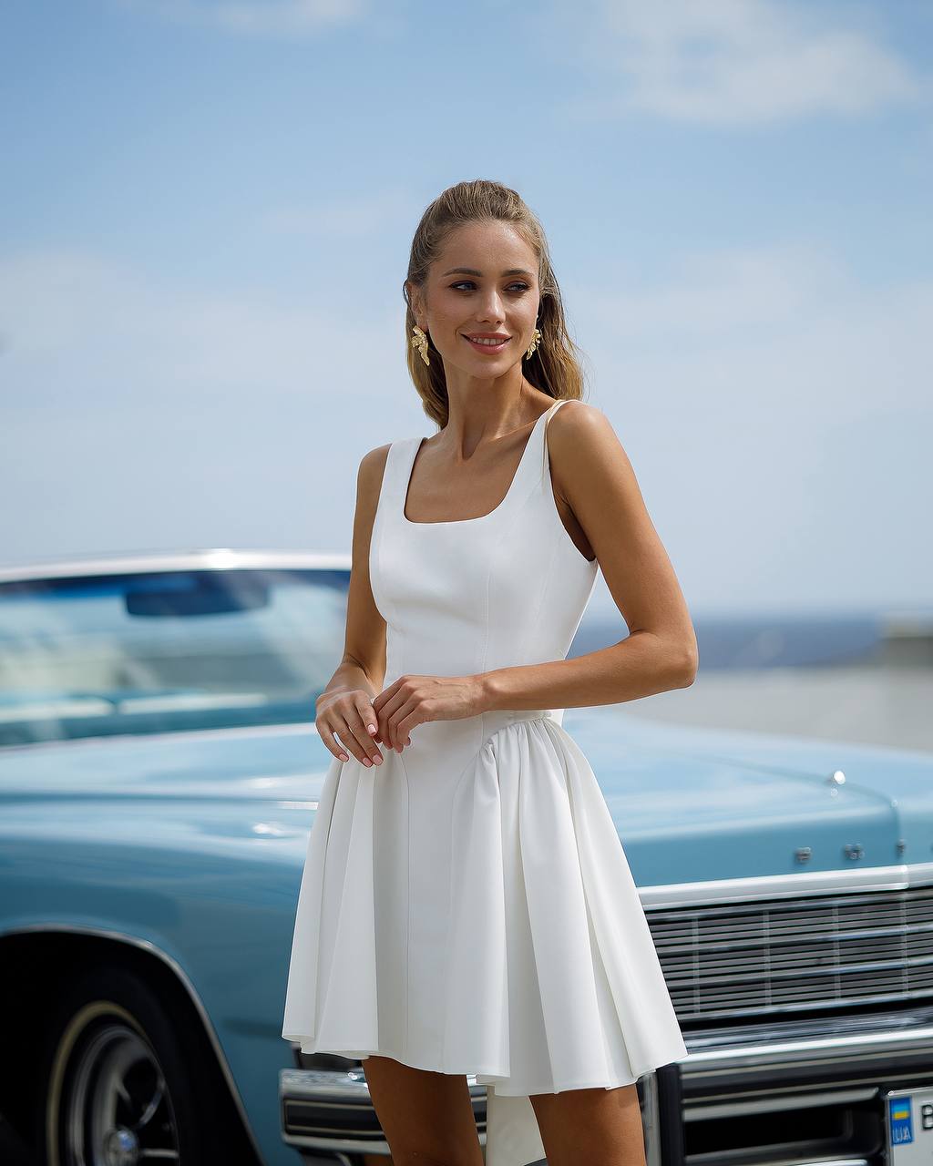 Woman in a white dress standing next to a vintage car with a clear sky background