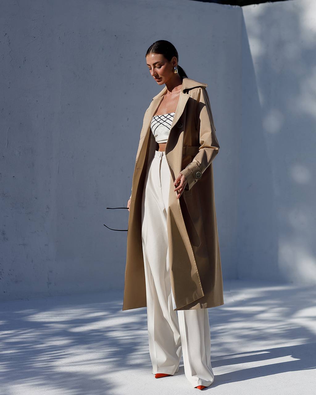 Woman wearing a beige trench coat, striped top, and white pants against a white wall.