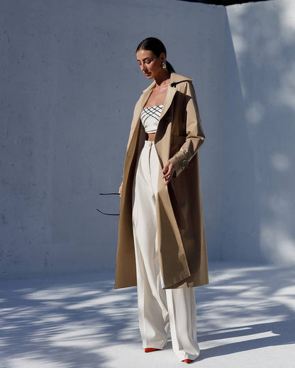 Woman wearing a beige trench coat, striped top, and white pants against a white wall.