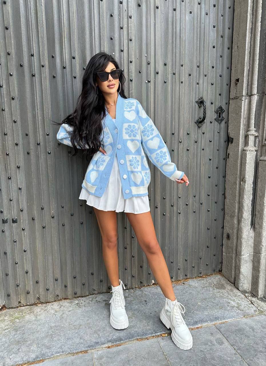 Woman wearing a light blue cardigan with a pattern, white skirt, and white sneakers in front of a metallic door.