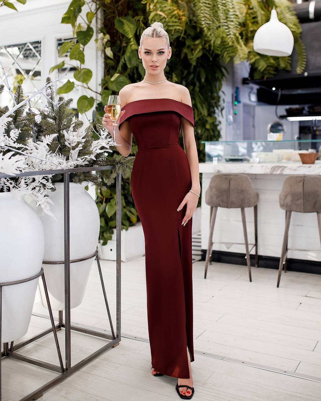 Woman in a burgundy off-shoulder dress holding a glass in an outdoor setting.