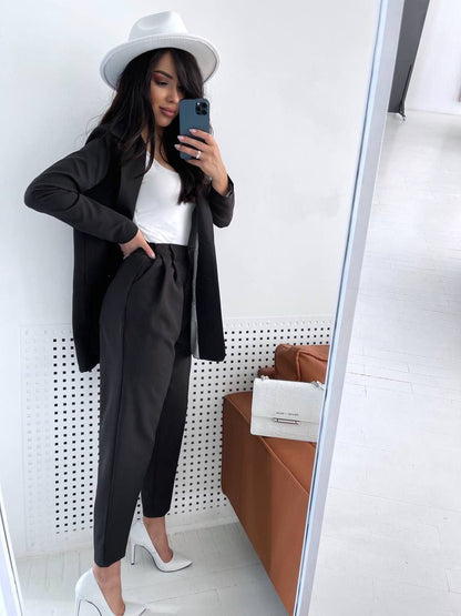 Woman taking a mirror selfie wearing a black blazer, white top, and pants with a white hat.