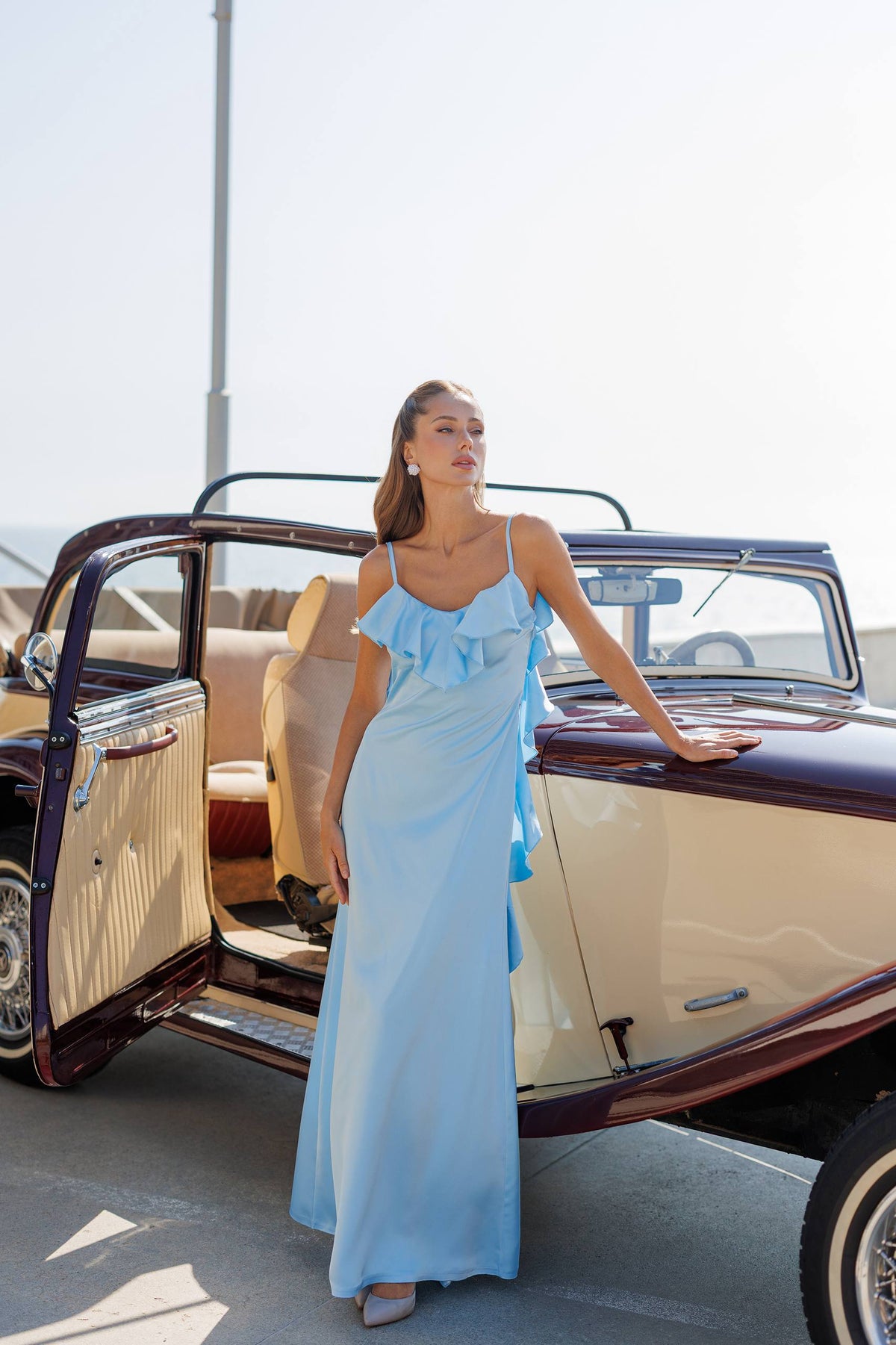 Woman in a light blue dress standing next to an old-fashioned car with open door.