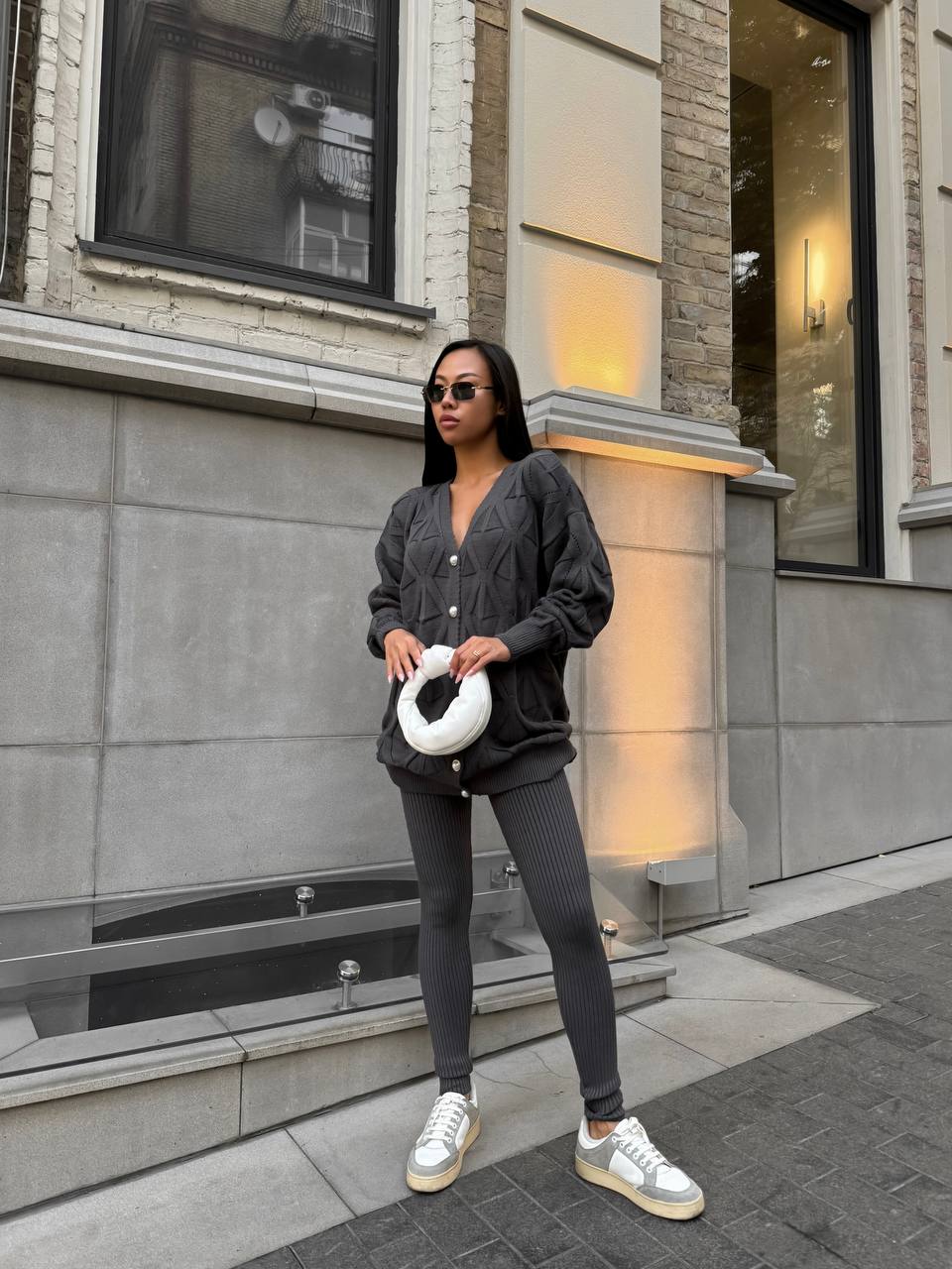 Woman standing outside a building wearing a dark gray outfit with white sneakers.