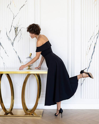 Woman in a black off-shoulder dress standing next to a gold table against a marble wall.