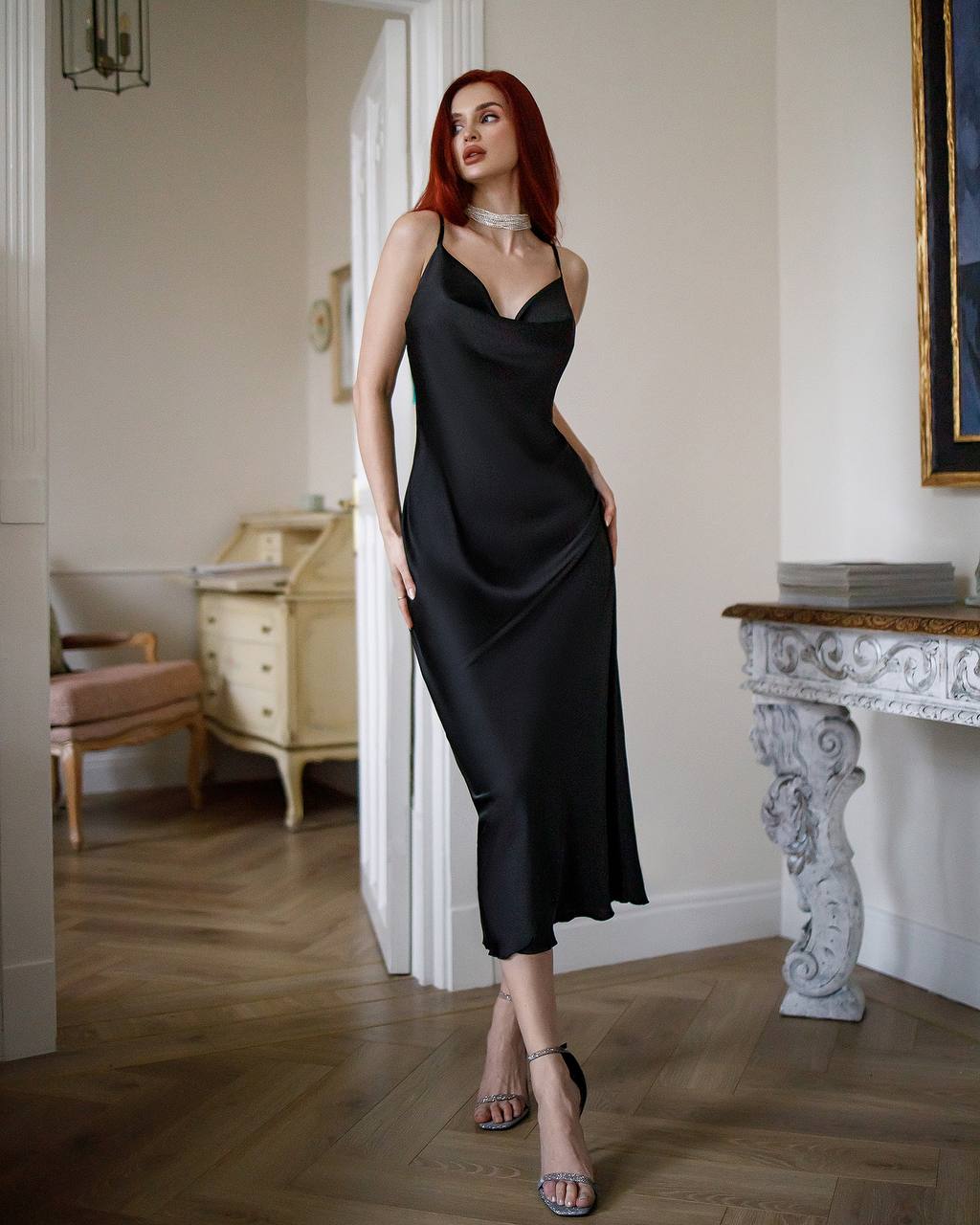 Woman in a black slip dress standing in an elegant room with wooden floors and white walls.