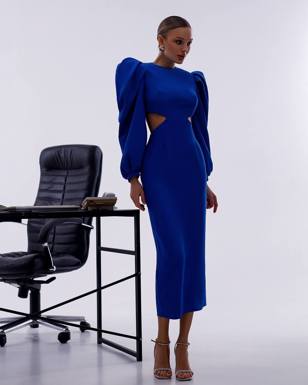 Woman in a blue dress standing next to a black office chair and desk on a white background