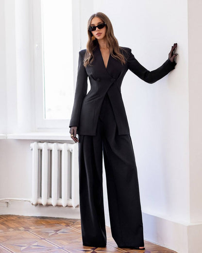 Woman in a black suit standing in a room with white walls and wooden floor.