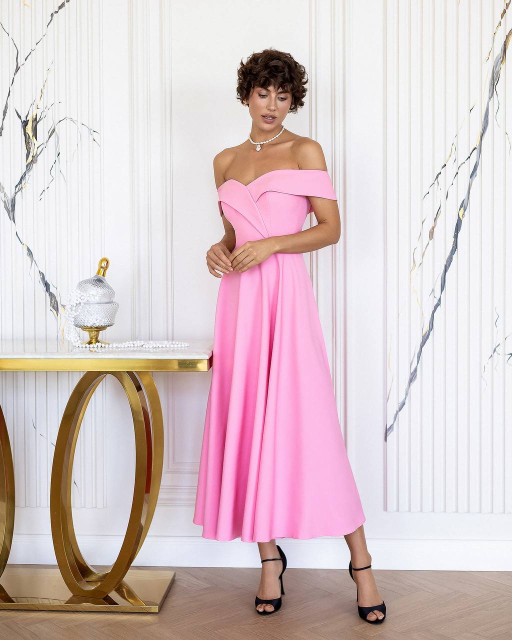 Woman in a pink off-shoulder dress standing in a stylish interior setting.
