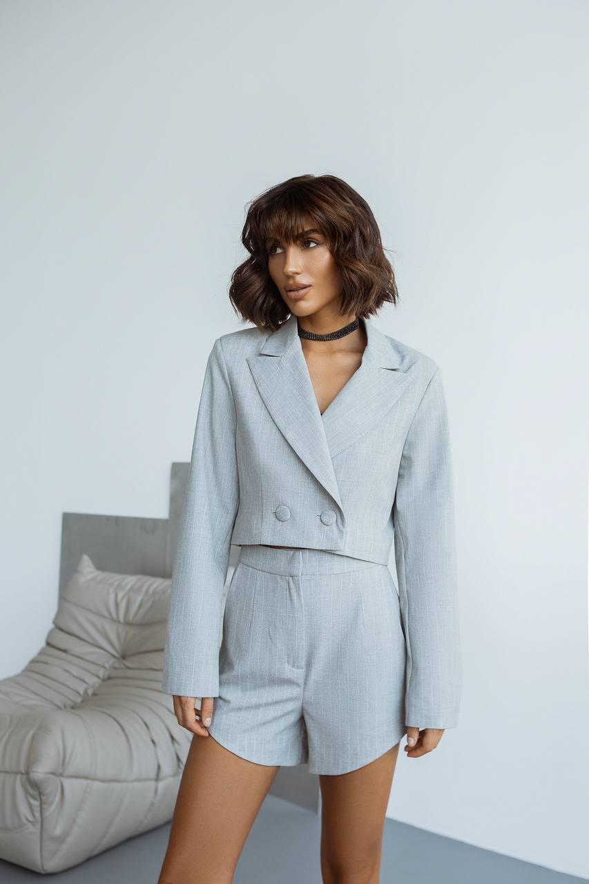 Woman wearing a light gray suit standing against a white wall with a gray chair.