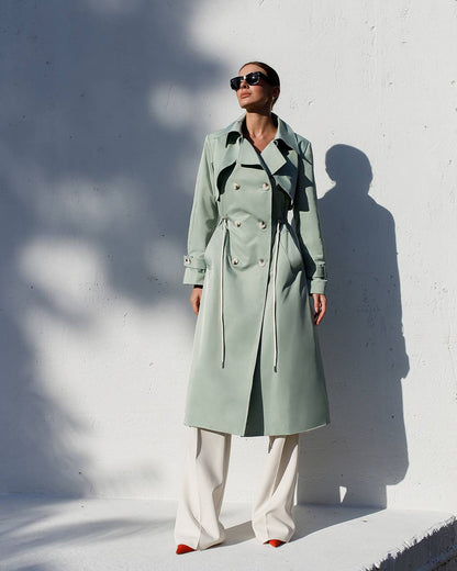 Person wearing a long light green coat against a white wall