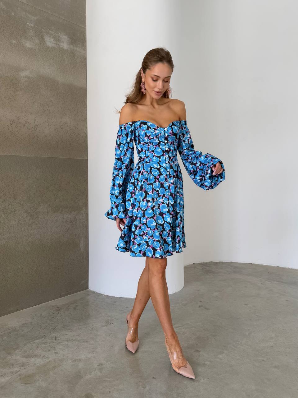 Woman wearing a blue floral off-shoulder dress against a plain wall.