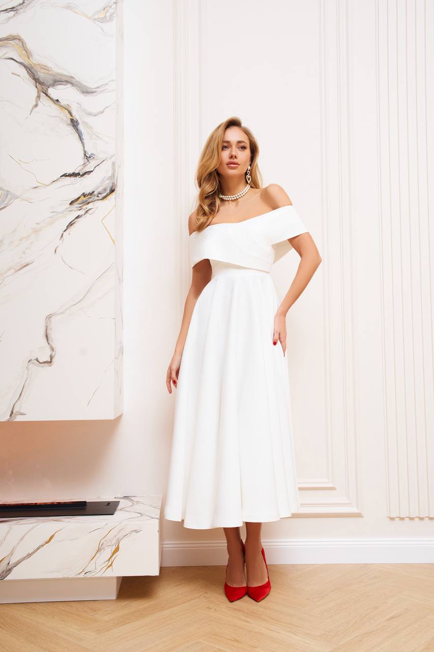 Woman in a white off-shoulder dress standing in a stylish interior setting.